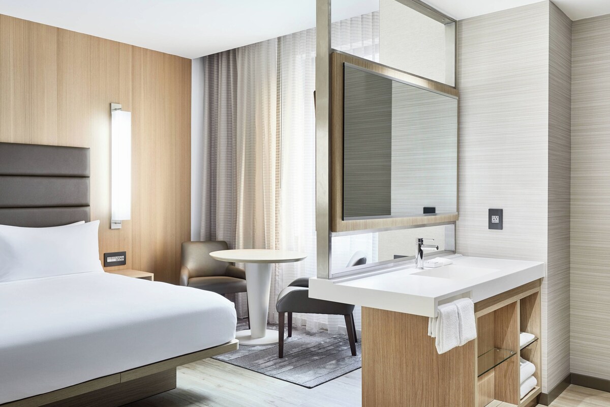 Una camera d'albergo moderna con un letto grande, un tavolo rotondo, un bagno con uno specchio ampio e un comò in legno.