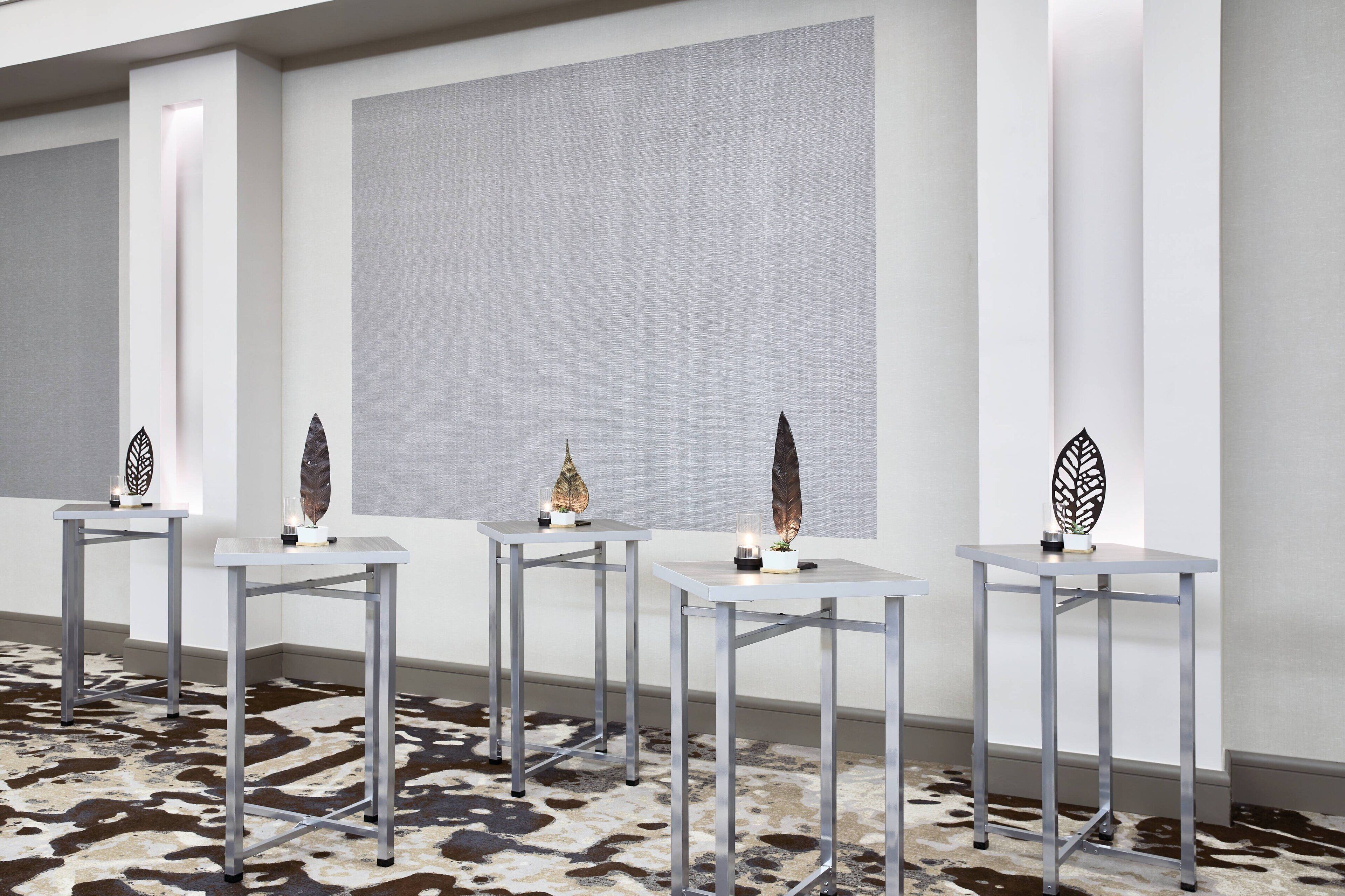 A modern interior with four white tables, each with a decorative object and candle. The room has a patterned carpet and a large grey wall.
