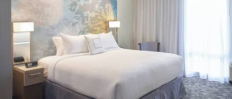 A neatly made bed with a white comforter and pillows, a bedside table with a lamp, and a patterned carpet.