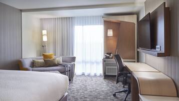 A hotel room with a large bed, a desk, a chair, a TV mounted on the wall, and a window with curtains.