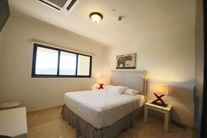 A bedroom with a large bed, two bedside tables with lamps, a window, and a painting of an elephant. - Aruba Stop Vacation Rentals (Noord)
