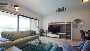 A modern living room with a sofa, a glass coffee table, a TV, and a bookshelf. - Aruba Stop Vacation Rentals (Noord)
