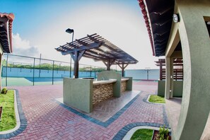 An outdoor patio area with a barbecue station, a pergola, and a view of a tennis court. - Aruba Stop Vacation Rentals (Noord)