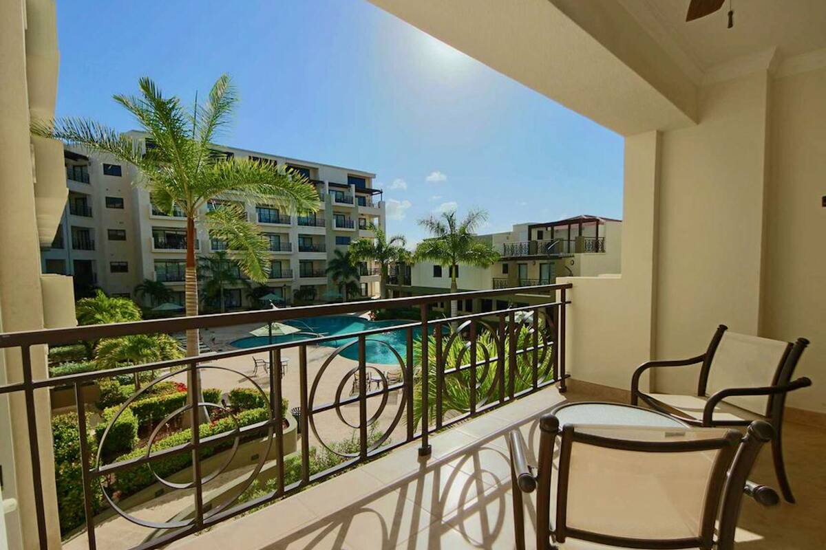 A balcony with a view of a swimming pool, palm trees, and a multi-story building in the background.
