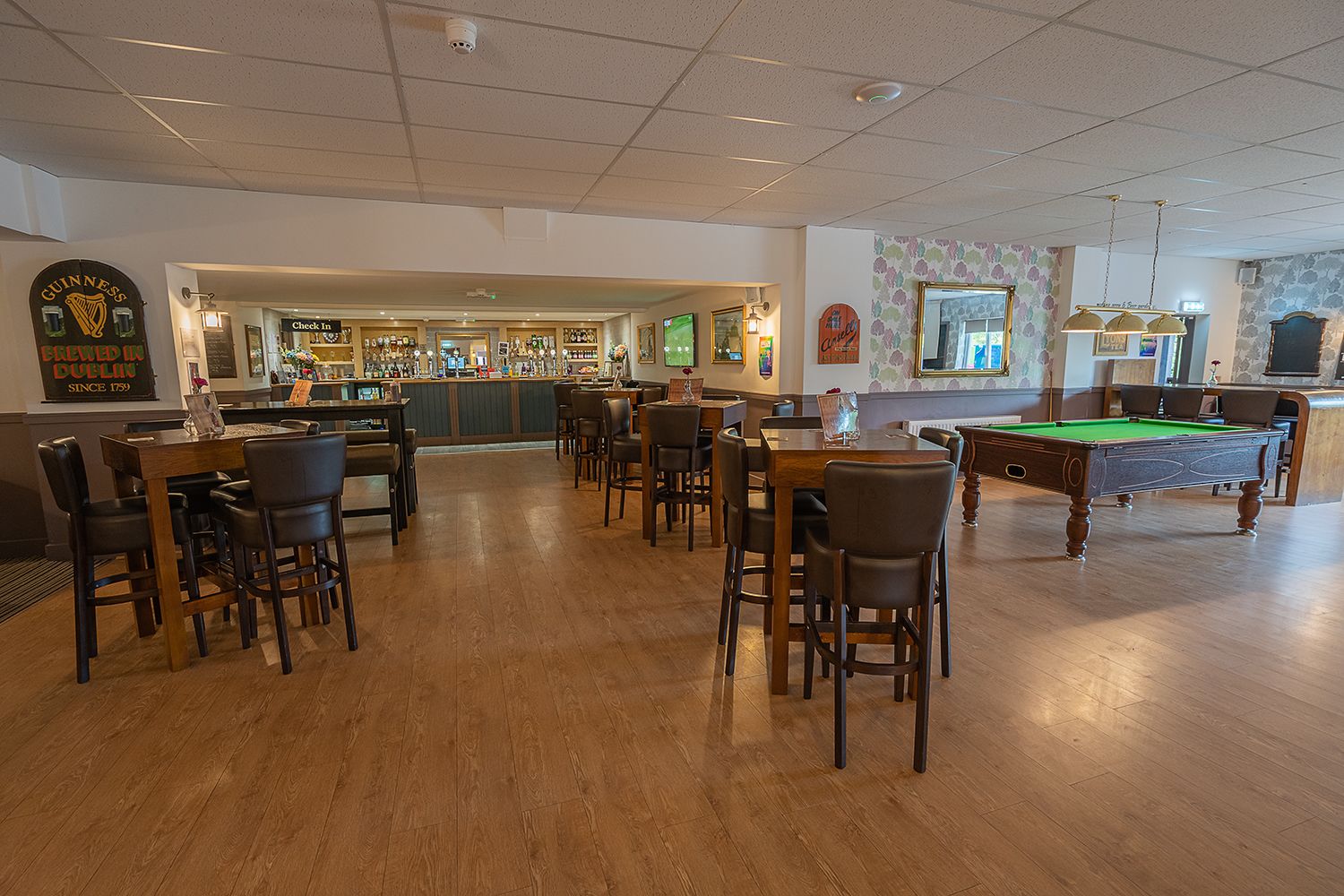 A bar area with a pool table, wooden flooring, and bar stools.