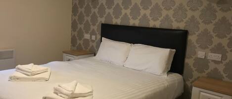 Una cama bien hecha con sábanas y almohadas blancas, una mesita de noche con lámpara, y una pared con papel tapiz estampado.
