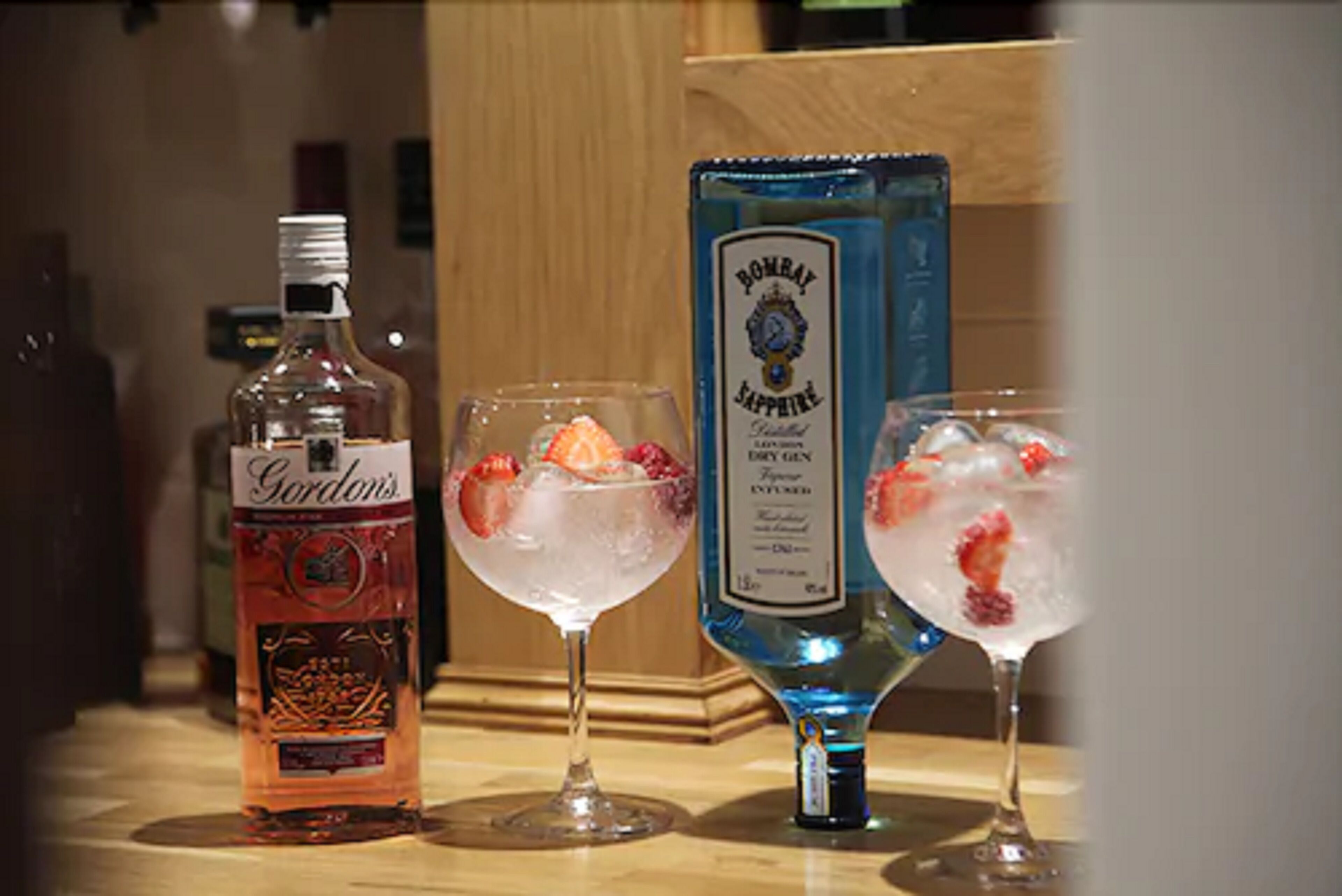 Una botella de ginebra Gordon's y dos vasos con hielo y fresas sobre una superficie de madera.