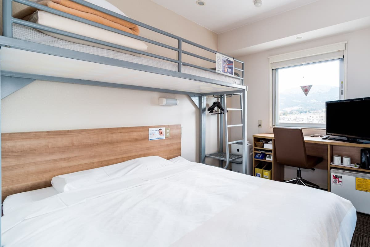 A hotel room with a bunk bed, a desk with a chair, a television, and a window with a view.