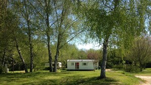 A single-story cabin with a red door, surrounded by trees and a dirt path. - Camping de Lagraulière (Perpezac-le-Noir)