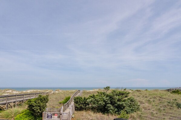 A coastal walkway leading to a beach with a wooden boardwalk and a sign indicating a beach access point.