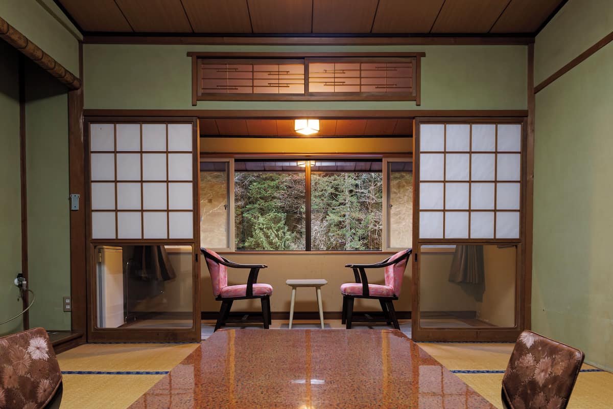 Une pièce de style japonais traditionnel, avec un sol recouvert de tatamis, une table basse, deux chaises et une fenêtre donnant sur un paysage verdoyant.