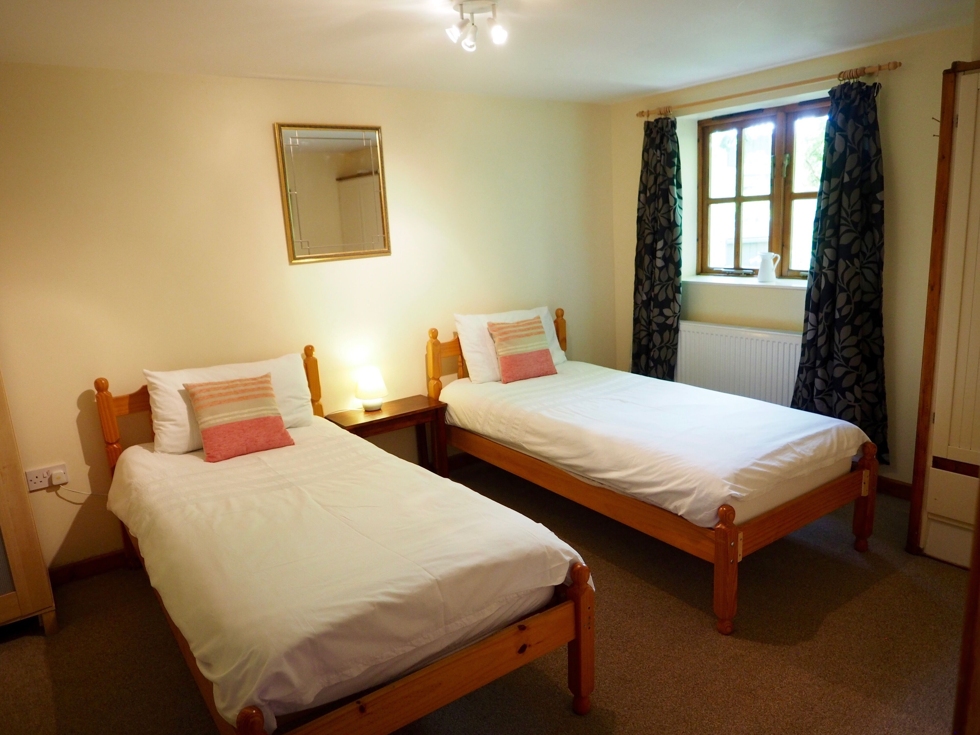 A room with two wooden beds, each with white bedding and pink pillows. There is a mirror on the wall above the beds, and a window with curtains to the right.