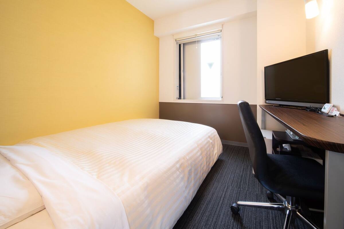 Ein Hotelzimmer mit einem Einzelbett, einem Schreibtisch mit Fernseher und einem Fenster mit Jalousien.