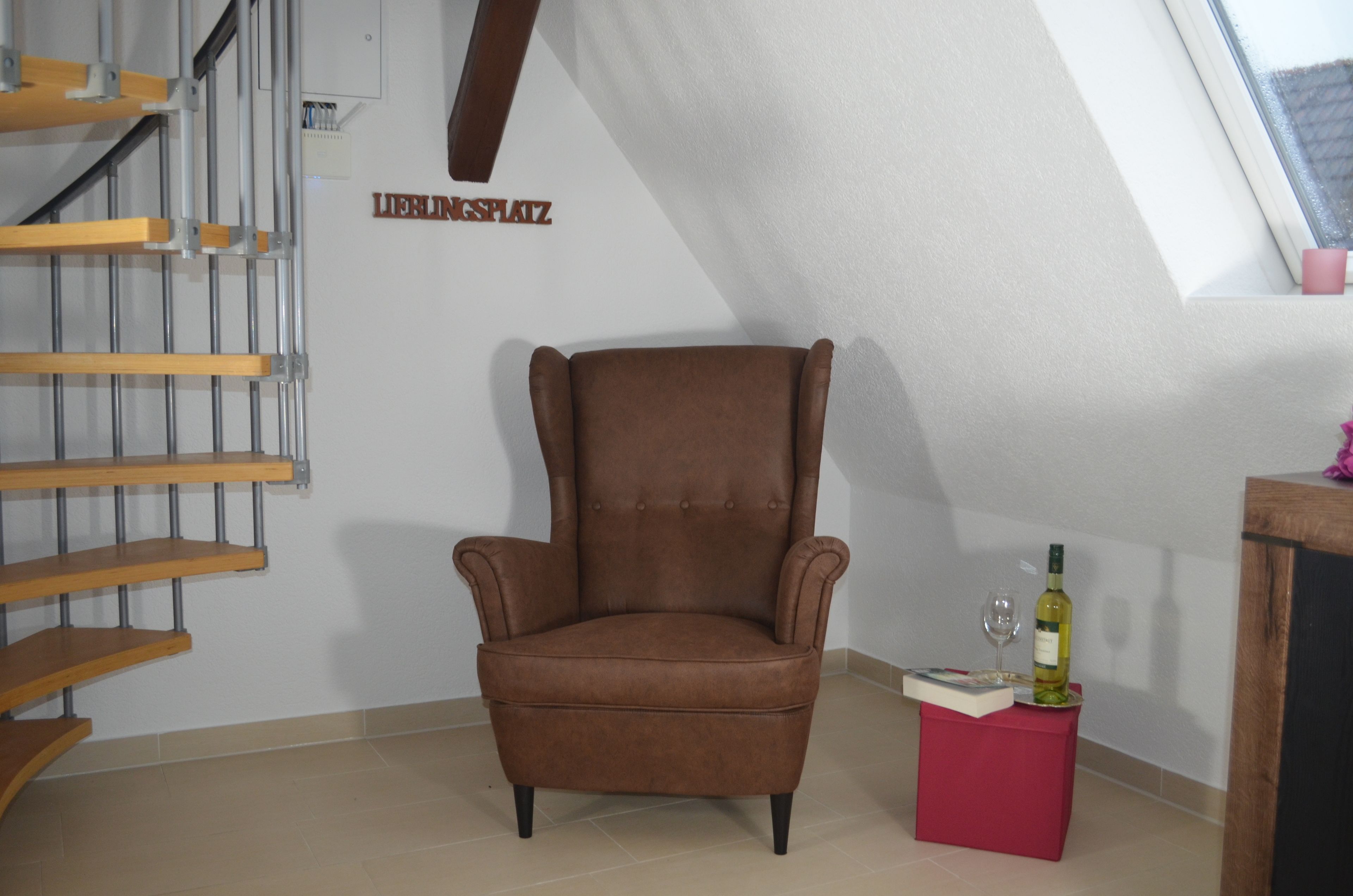 A brown armchair in a room with a sloped ceiling, a wooden staircase, and a small red table with a wine bottle and glass.