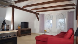 A living room with a red sofa, a TV on a wooden stand, a kitchen area with a stove, and a glass table with a vase. - Wolke65 (Naumburg)