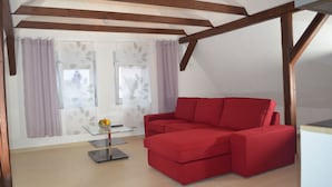 A red sofa in a room with a sloped ceiling and wooden beams, a glass table with a vase, and a window with purple curtains. - Wolke65 (Naumburg)