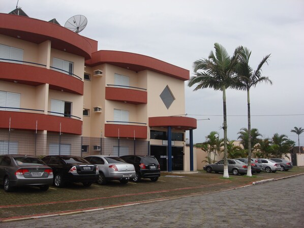 A multi-story building with a beige facade, balconies, and a parking lot with several cars.