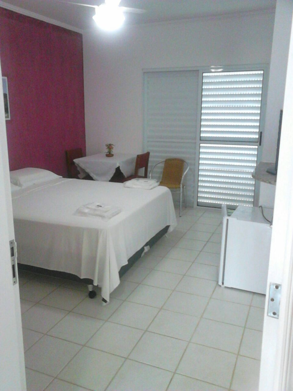 Habitaci ón de hotel con cama, una mesita con silla, ventana con persianas y pared blanca.