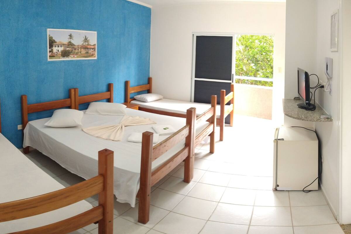 Une chambre d’hôtel avec deux lits simples, une tête de lit en bois, un petit bureau avec un ordinateur et un balcon donnant sur un paysage verdoyant.