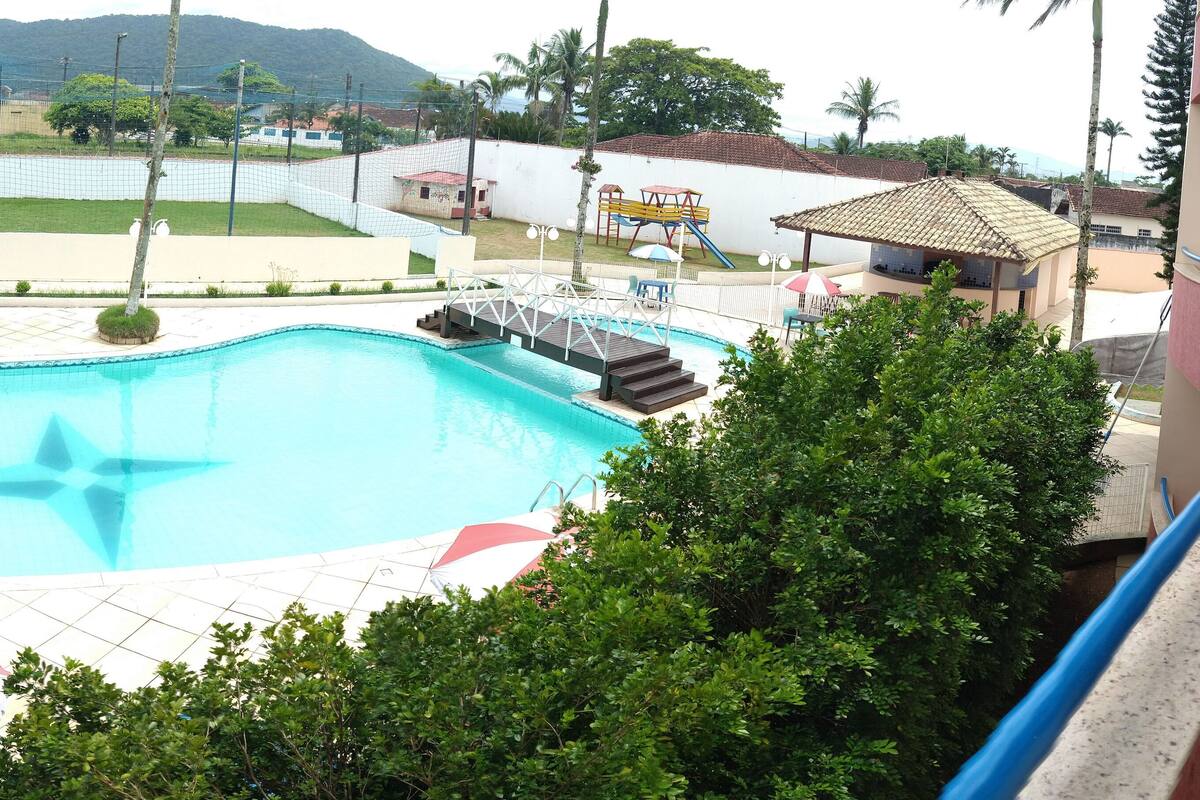 Une piscine de forme étoilée, entourée d’une clôture, avec une aire de jeux et un petit pavillon en arrière-plan.