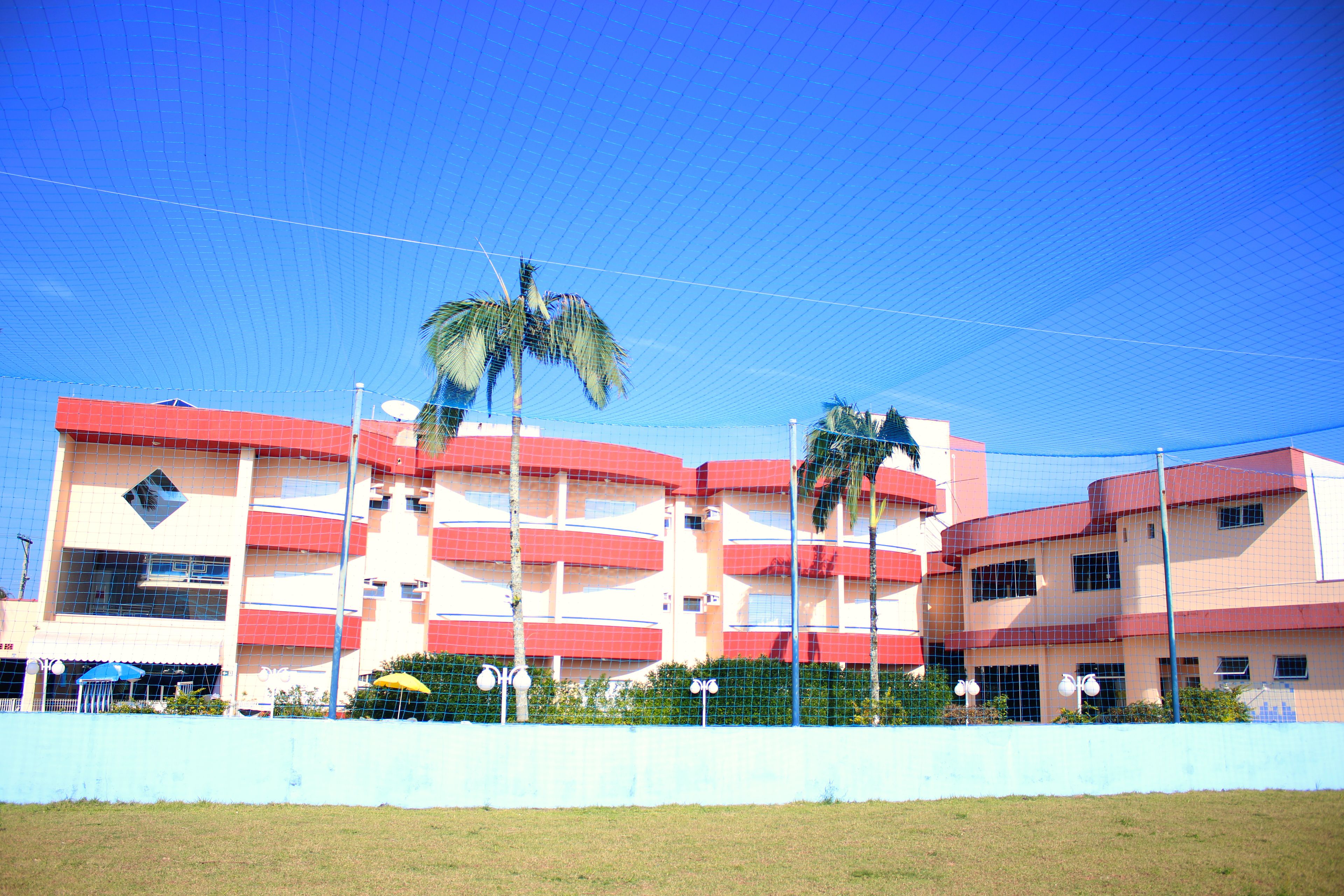 Un edificio de varios pisos con fachada roja y color crema, varios balcones y un campo deportivo cercado.