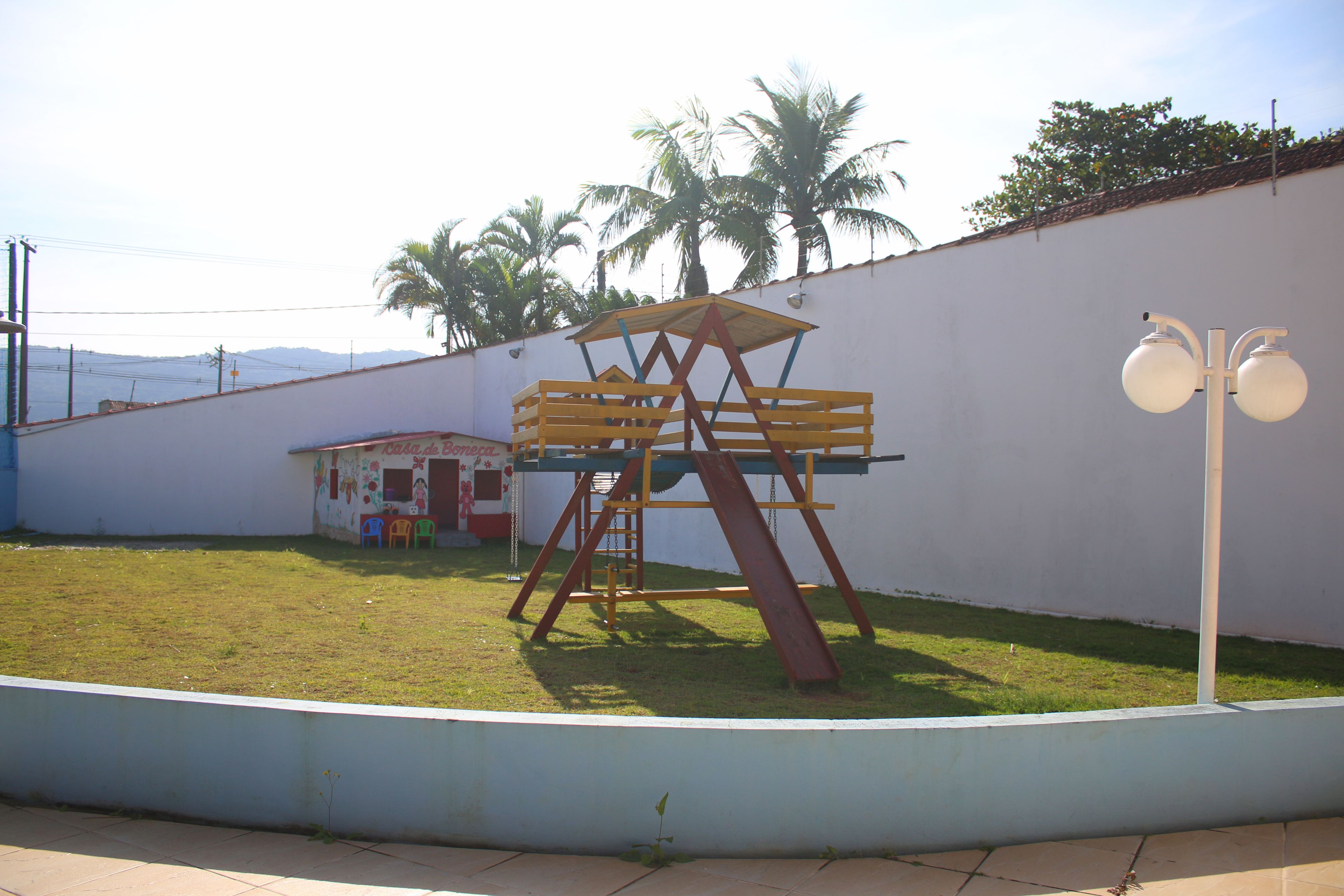 Um parquinho com escorregador e balanços, um pequeno edifício e palmeiras ao fundo.