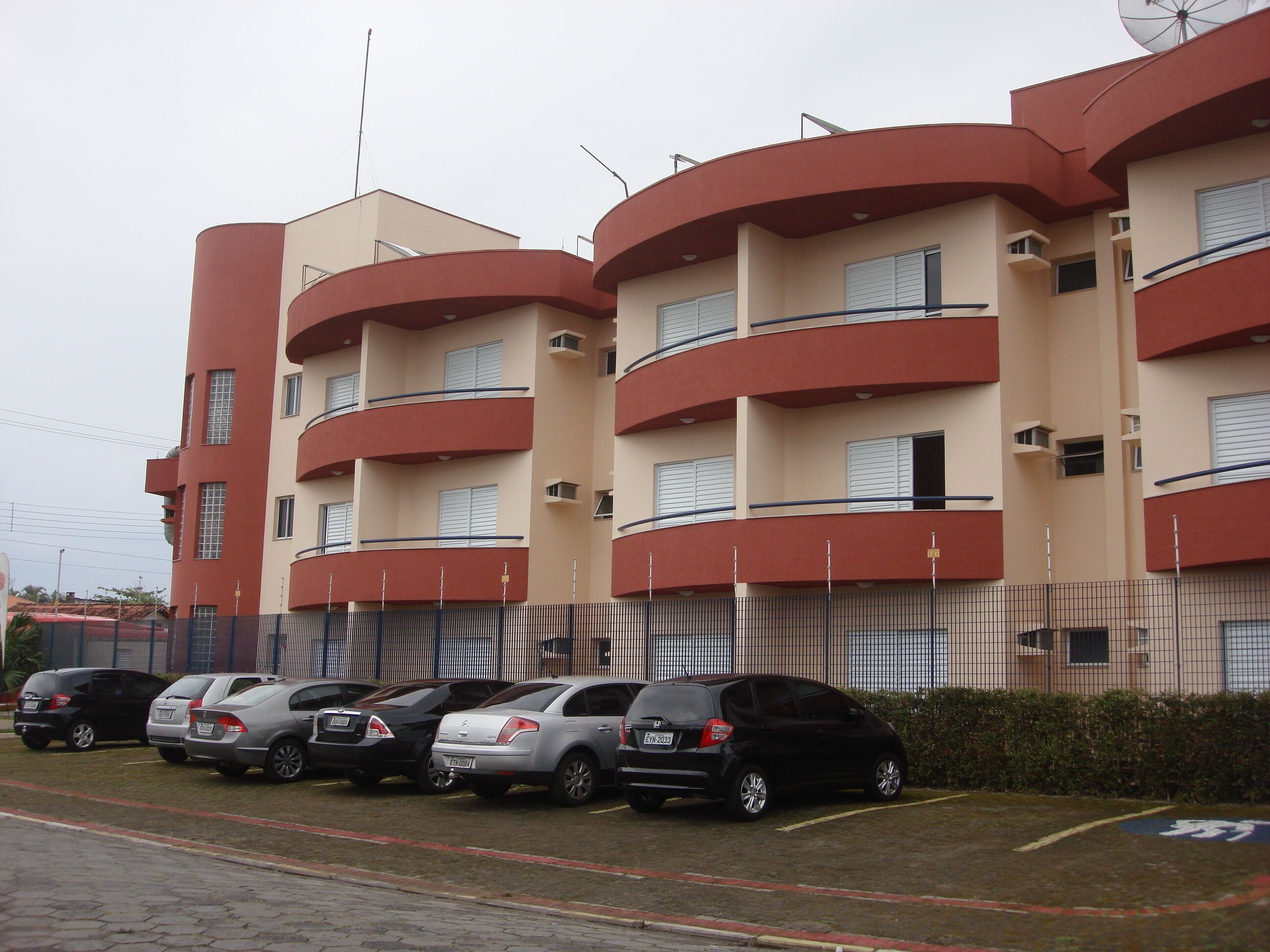 A multi-story residential building with balconies, a parking lot with several cars, and a fence.