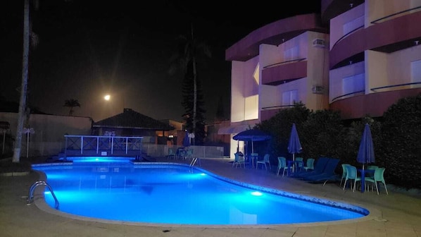 Un área de piscina nocturna con una luna de fondo, rodeada por un edificio de varios pisos con balcones.
