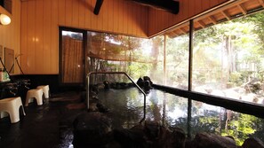 Un onsen japonés tradicional con interior de madera, un manantial termal de piedra y vistas a un área boscosa a través de amplios ventanales.