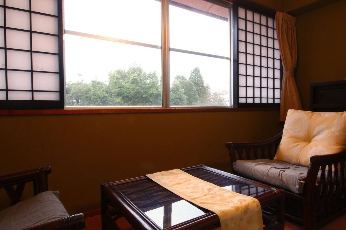 Een traditionele kamer met tatami vloerbedekking, een lage houten tafel, een comfortabele stoel en een groot raam met shoji schermen.
