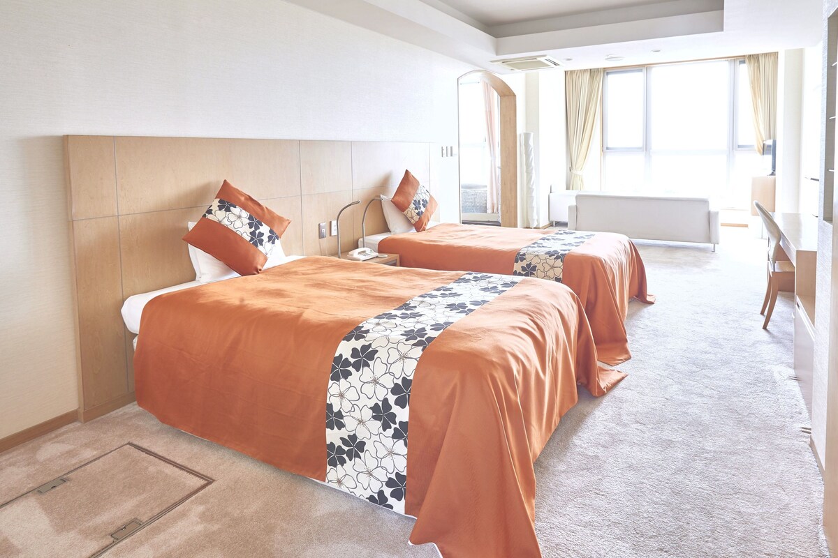 A hotel room with two beds, a desk, and a large window.