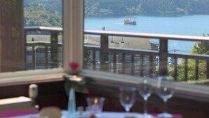 Una mesa de comedor con platos y vasos, una ventana con vistas a un lago y colinas, y una barandilla de balcón.