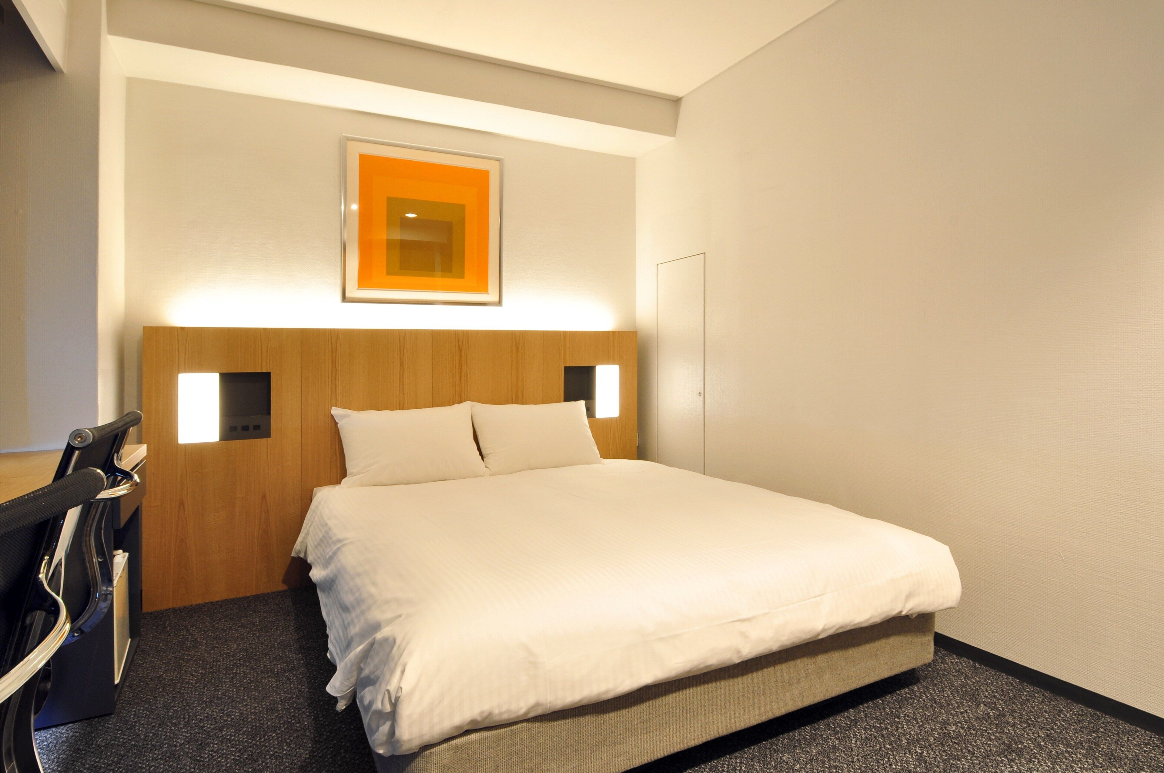 Ein Hotelzimmer mit Bett, Kopfteil, Wandleuchte und kleinem Schreibtisch mit Stuhl.