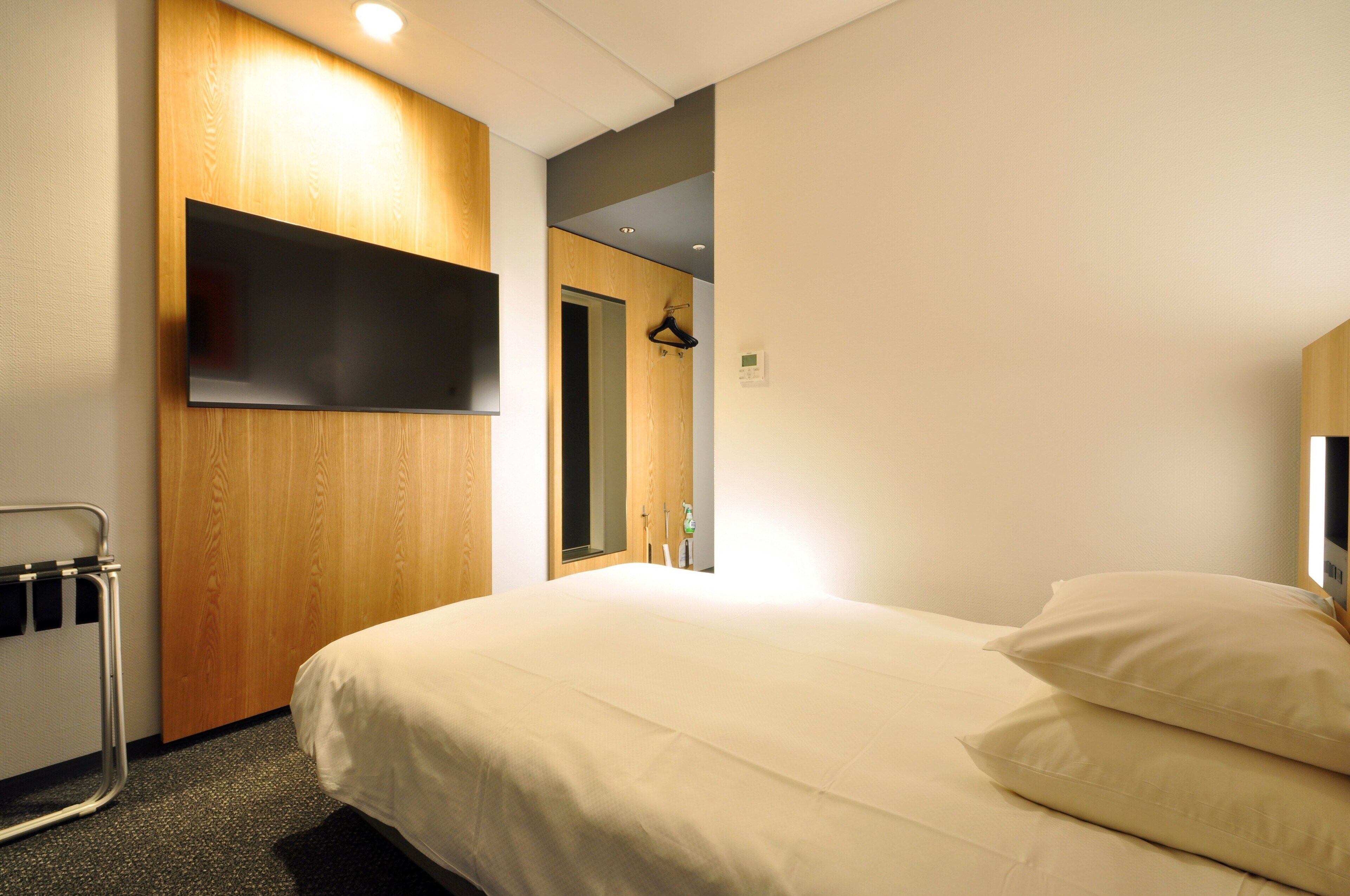 A hotel room with a bed, a TV mounted on the wall, a coat rack, and a door leading to another room.