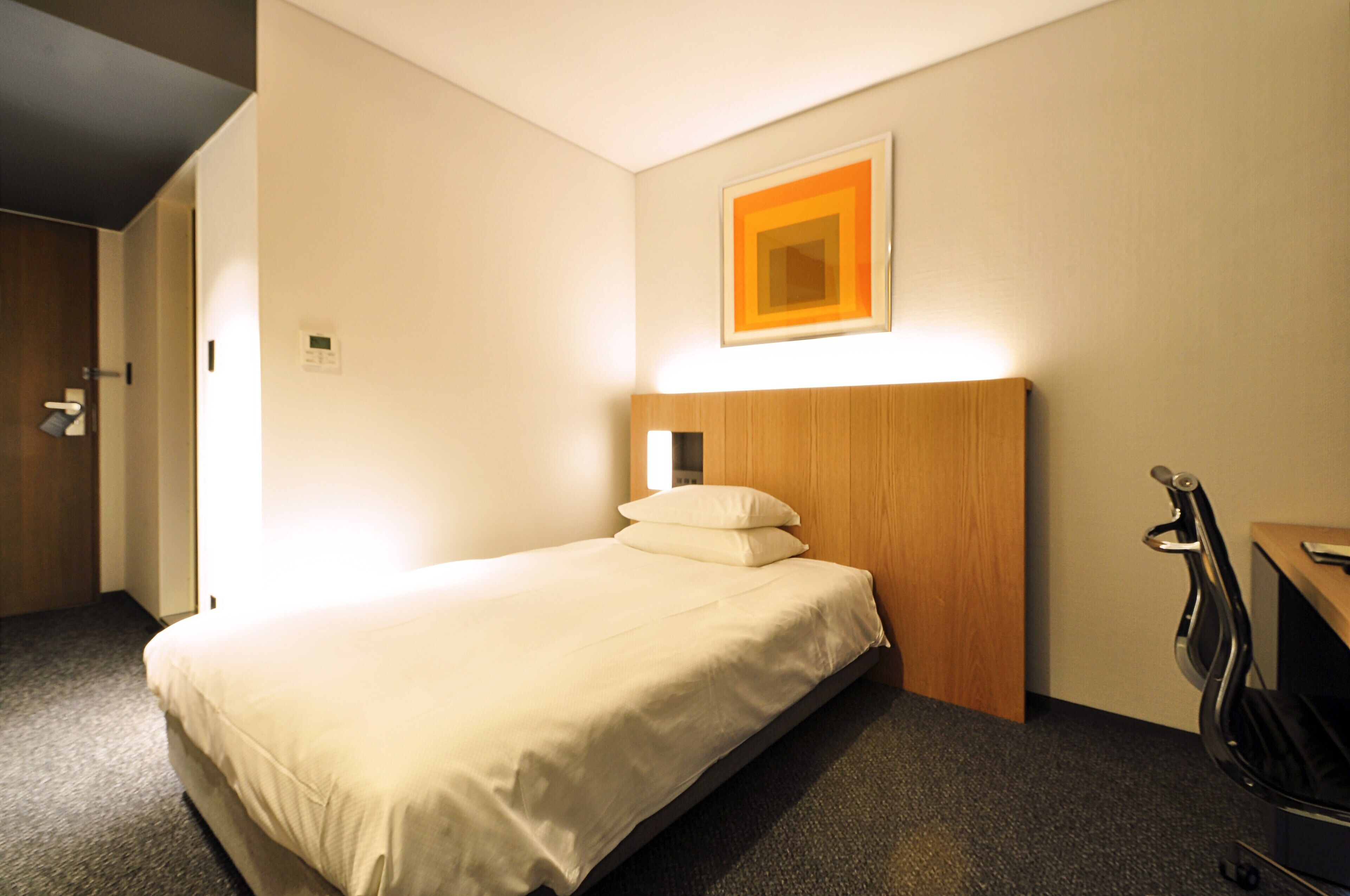 A hotel room with a bed, a wooden headboard, a desk with a chair, and a wall-mounted light fixture.