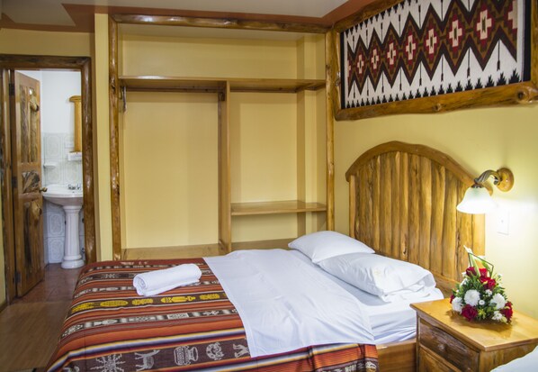 A hotel room with a bed, a wooden headboard, a nightstand with a lamp, a floral arrangement, and a wardrobe. - Hostal Santa Fe 1 (Otavalo)