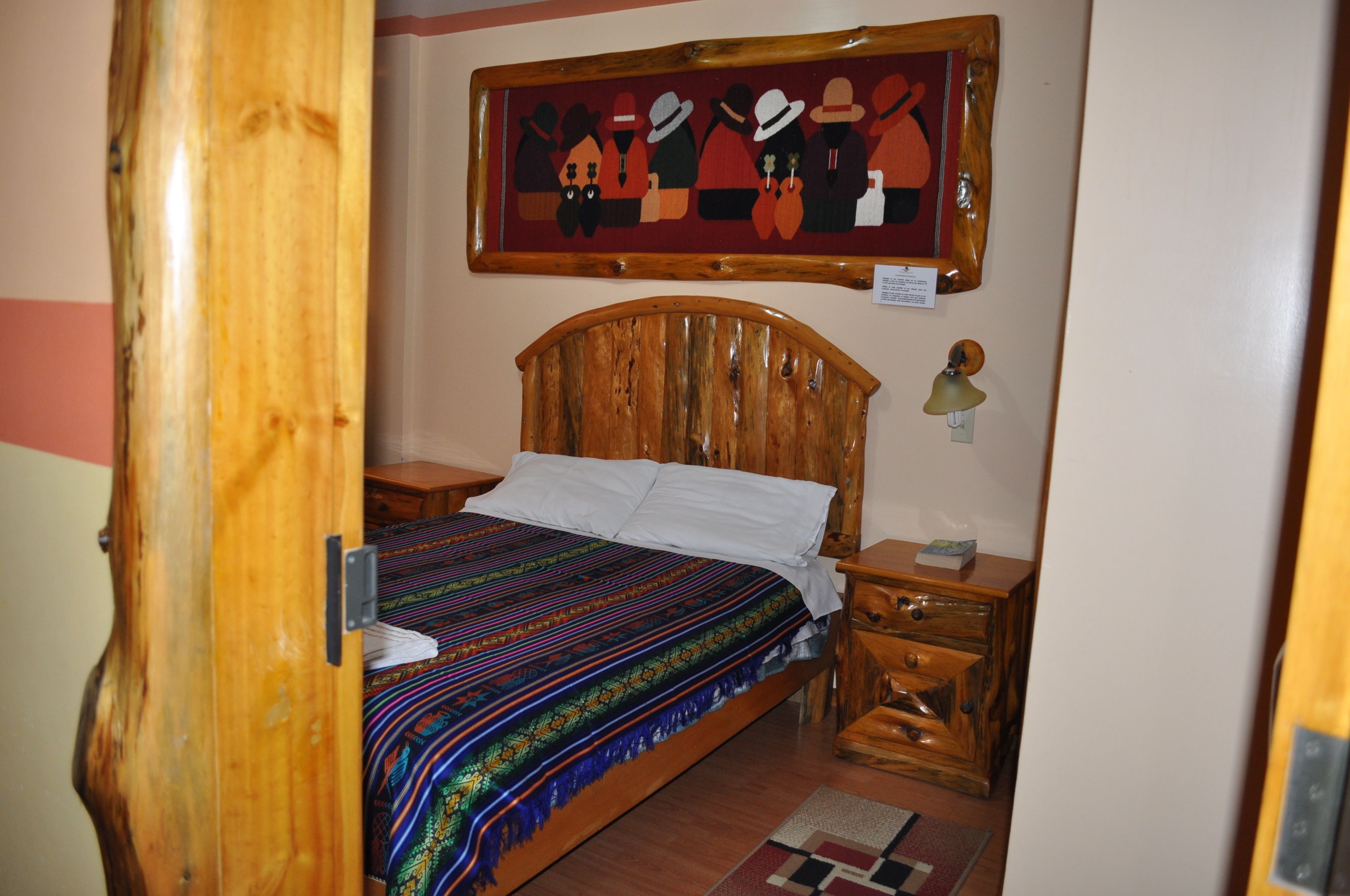 Una cama con estructura de madera, con un edredón colorido, una mesita de noche con una lámpara y un cuadro en la pared.