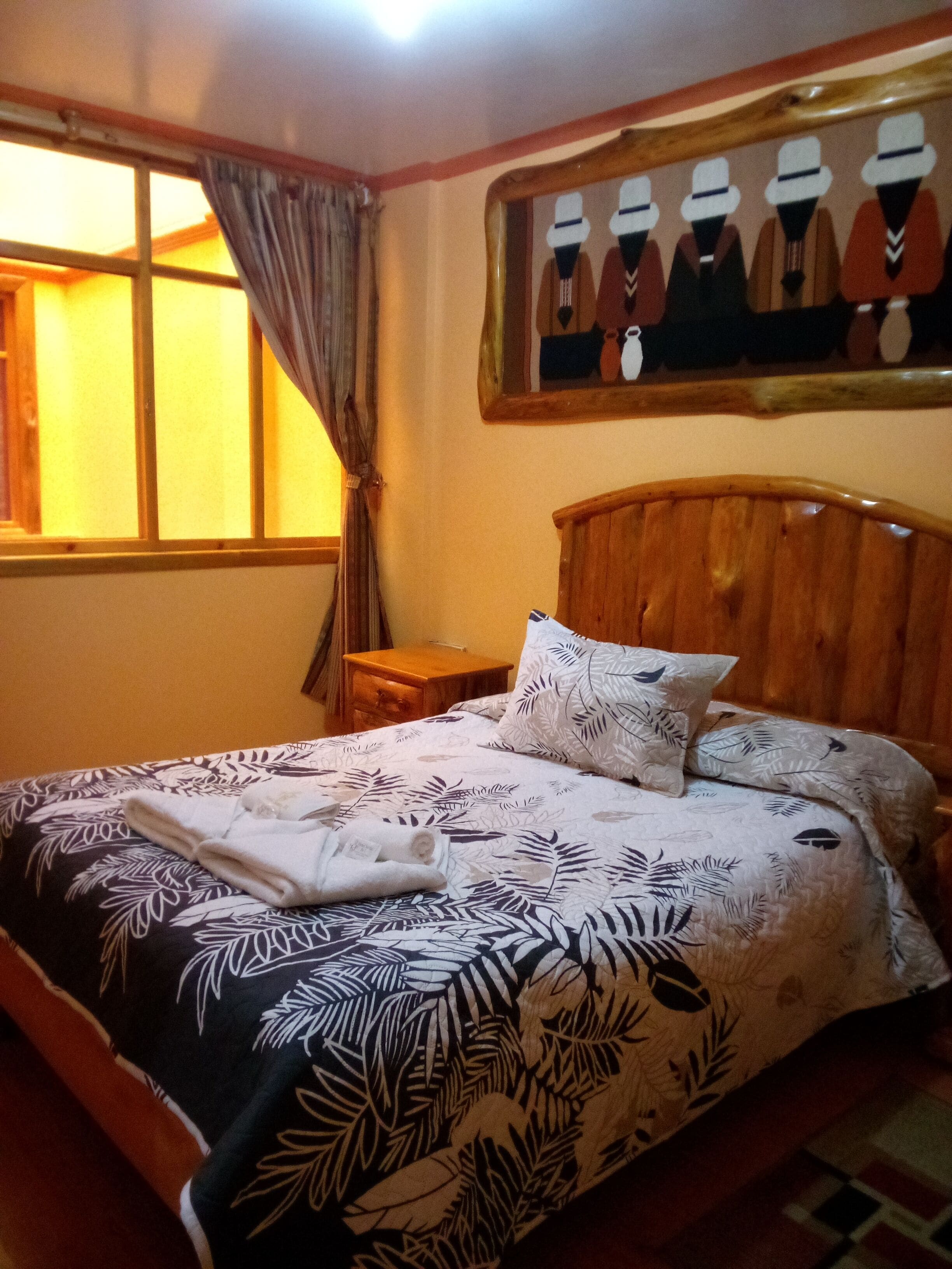 Un lit bien fait, recouvert d’une couvre-lit à motifs de feuilles, avec une tête de lit en bois et une table de chevet surmontée d’une lampe.