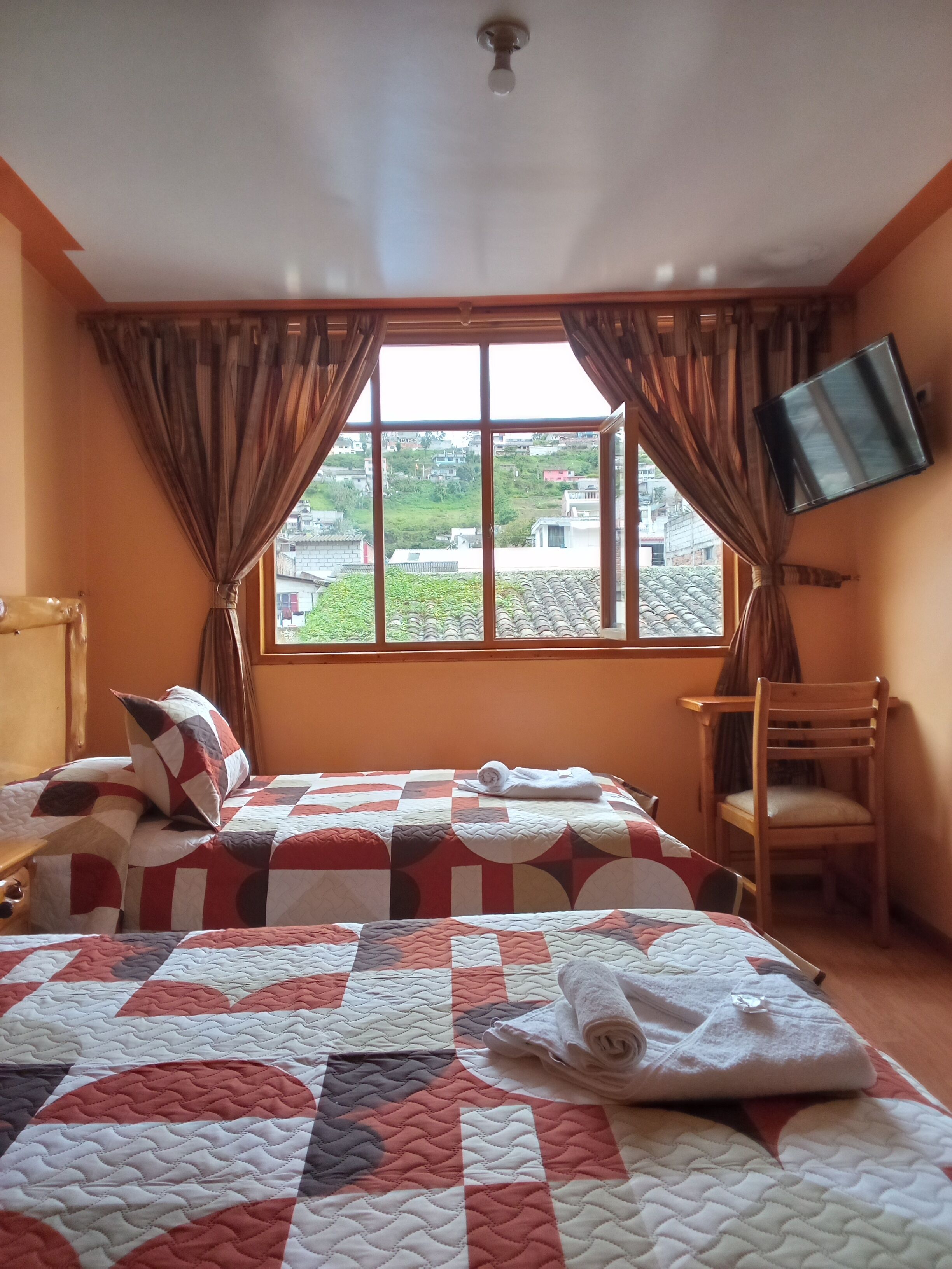 A hotel room with a large bed, a wooden chair, a flat-screen TV mounted on the wall, and a window with curtains.