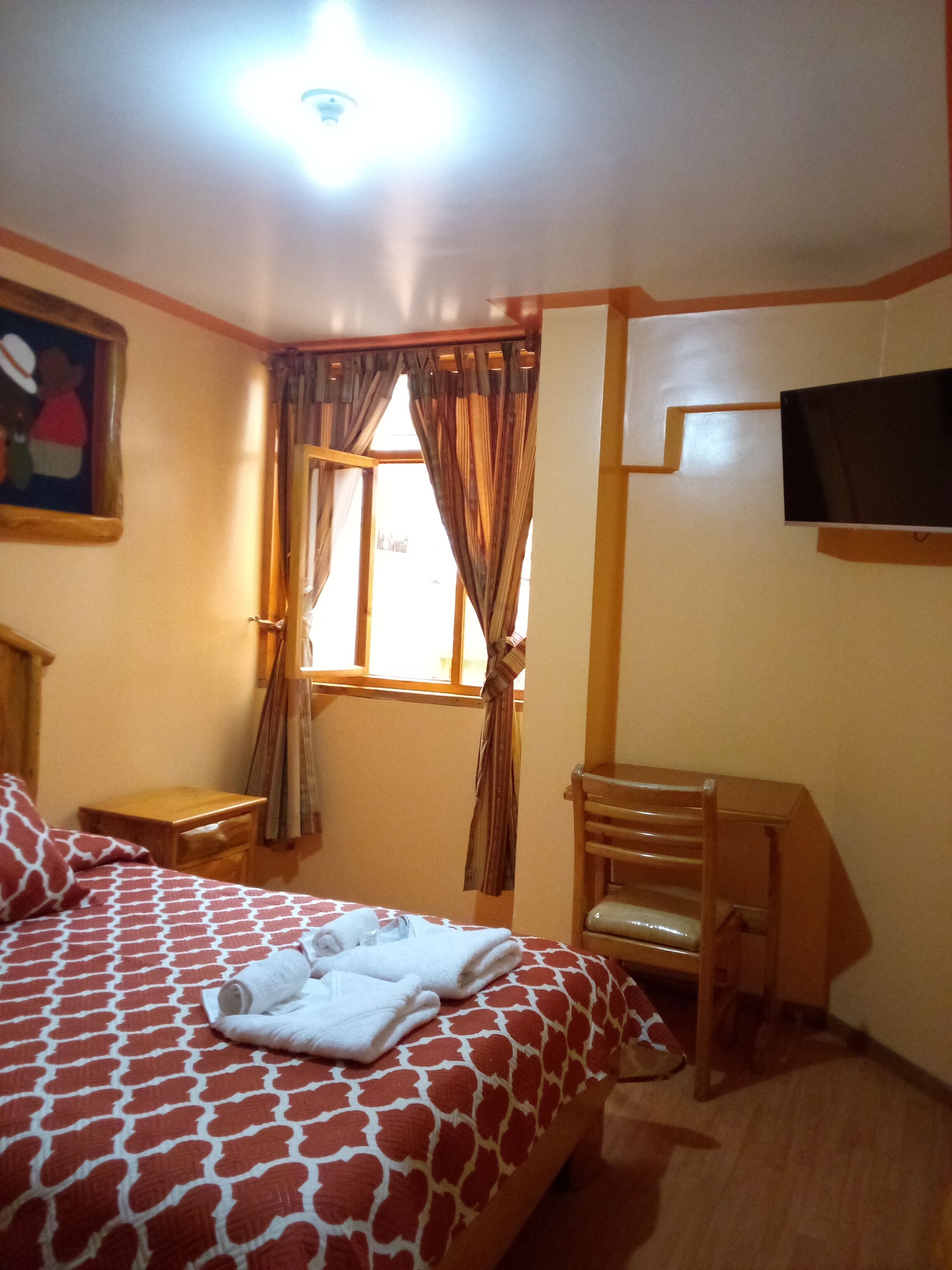 Habitación de hotel con cama, una silla, una mesita, televisor montado en la pared y una ventana con cortinas.
