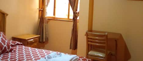 Habitación de hotel con cama, una silla, una mesita, televisor montado en la pared y una ventana con cortinas.