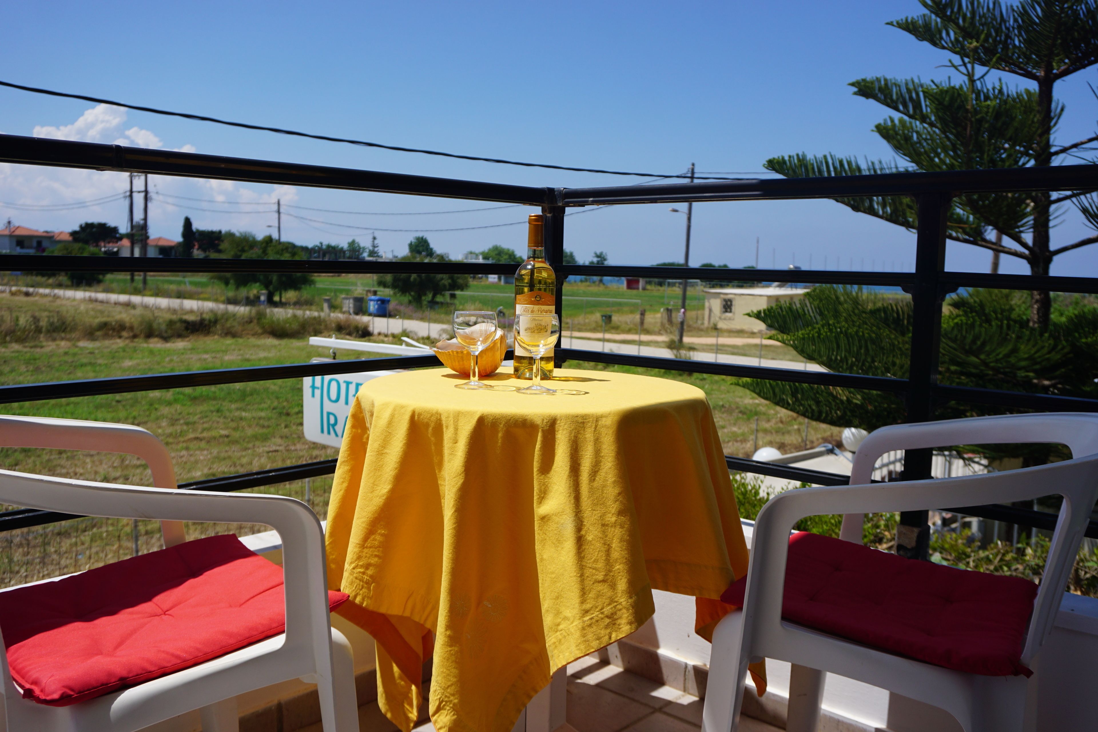 Une table recouverte d’une nappe jaune, deux chaises blanches avec des coussins rouges, une bouteille de vin et un verre de bière.