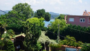 Ein üppiger Garten mit einem hölzernen Gartenpavillon, Blick auf einen See und einem Gebäude mit rosa Fassade und Ziegeldach.