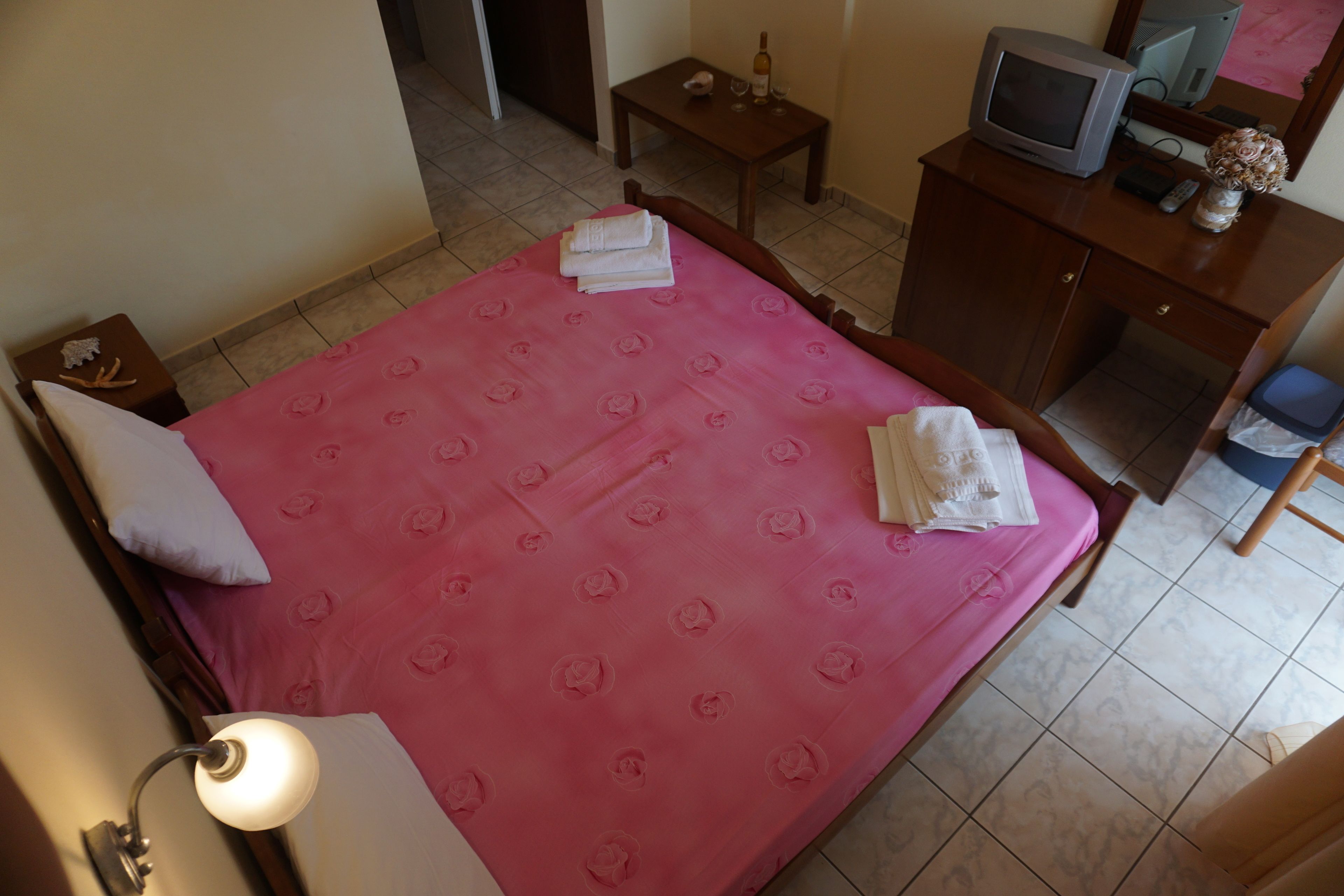 Une chambre d’hôtel avec un grand lit, un téléviseur posé sur une commode en bois, une petite table avec une bouteille et des verres, et une lampe fixée au mur.