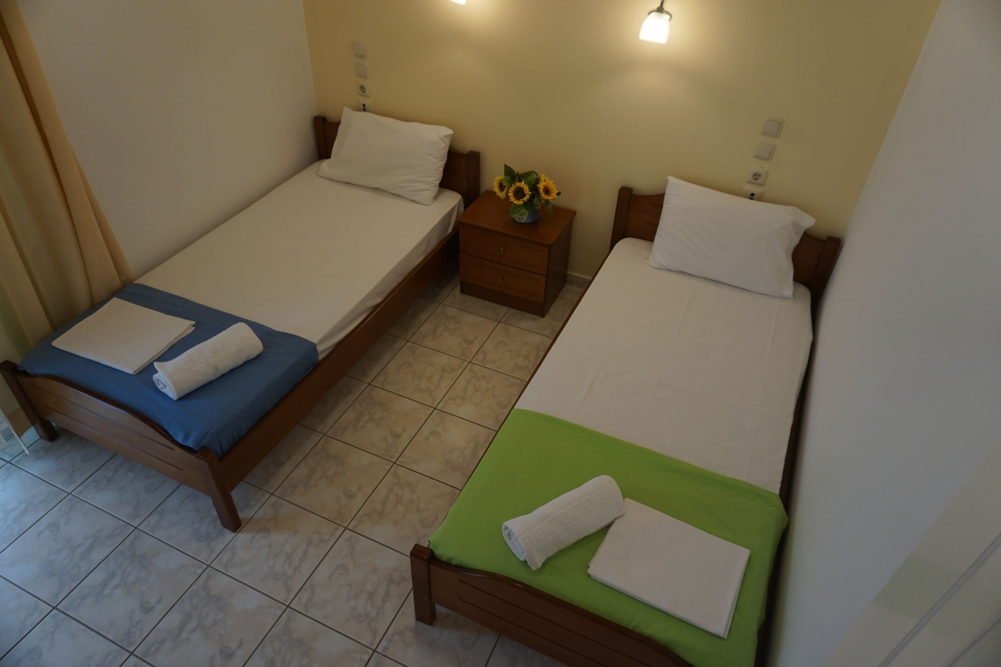 A hotel room with two single beds, a small wooden nightstand with a vase of sunflowers, and a green and blue bedspread.
