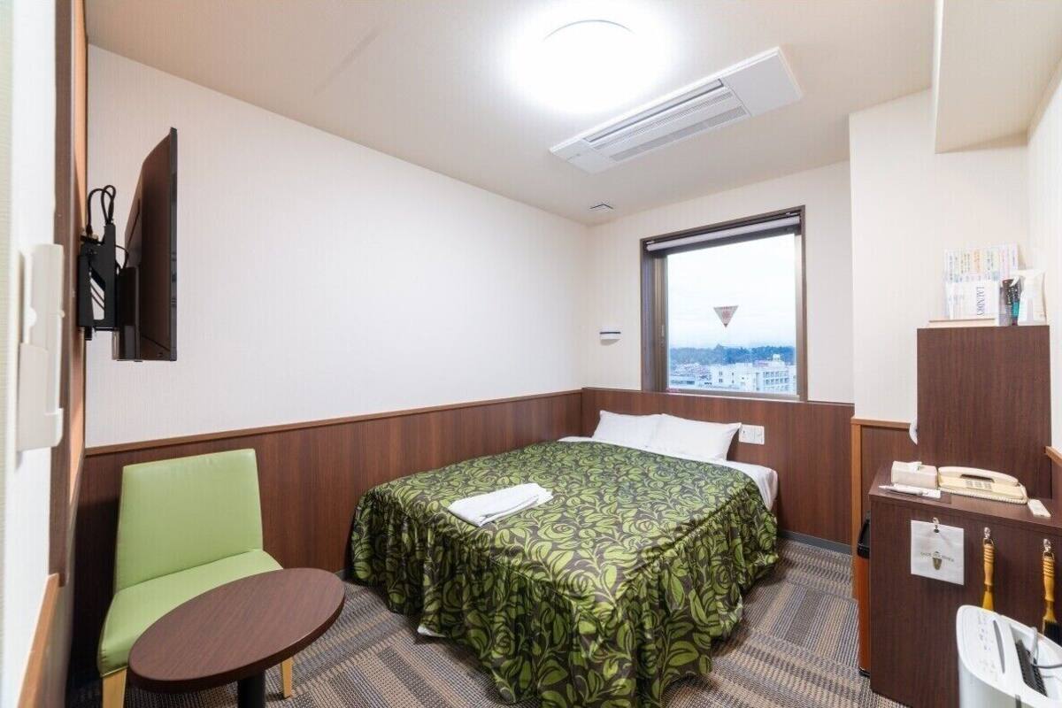 A hotel room with a bed, a green chair, a small table, a desk with a telephone, and a window with a view.
