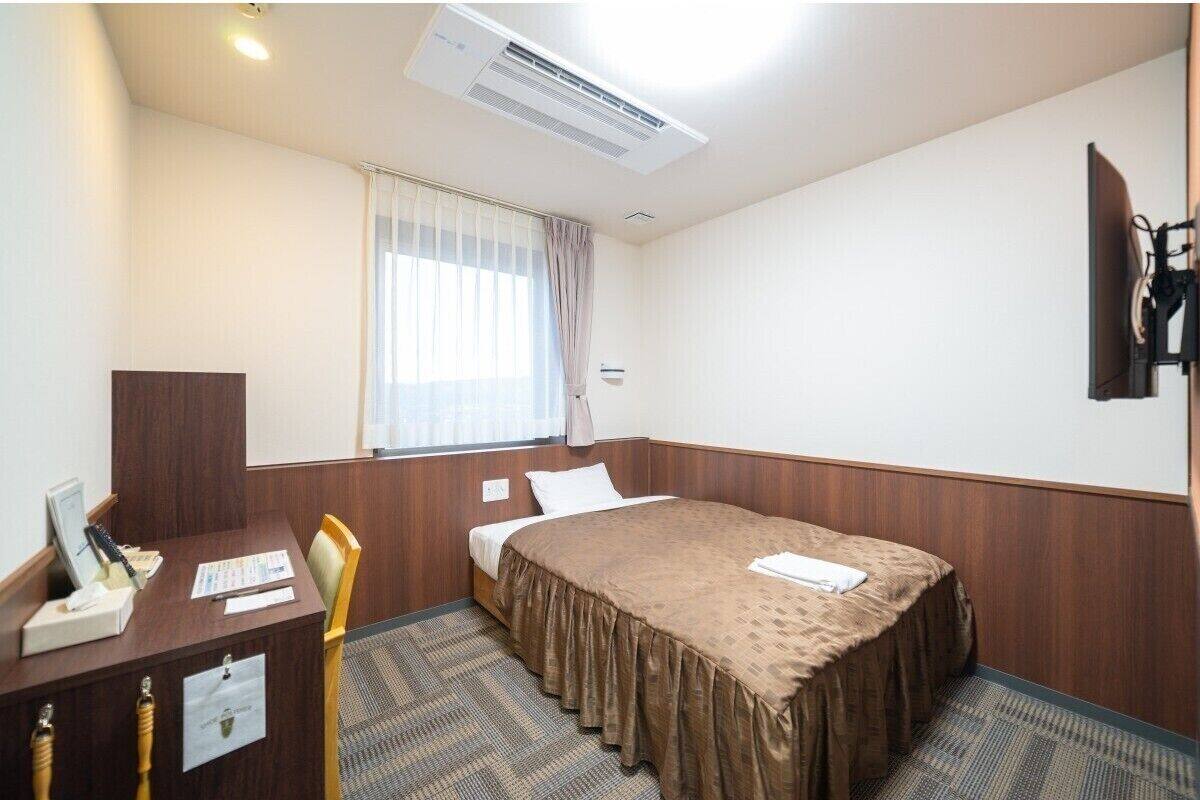 A hotel room with a bed, a desk with a chair, a window with curtains, an air conditioning unit, and a flat-screen TV mounted on the wall.