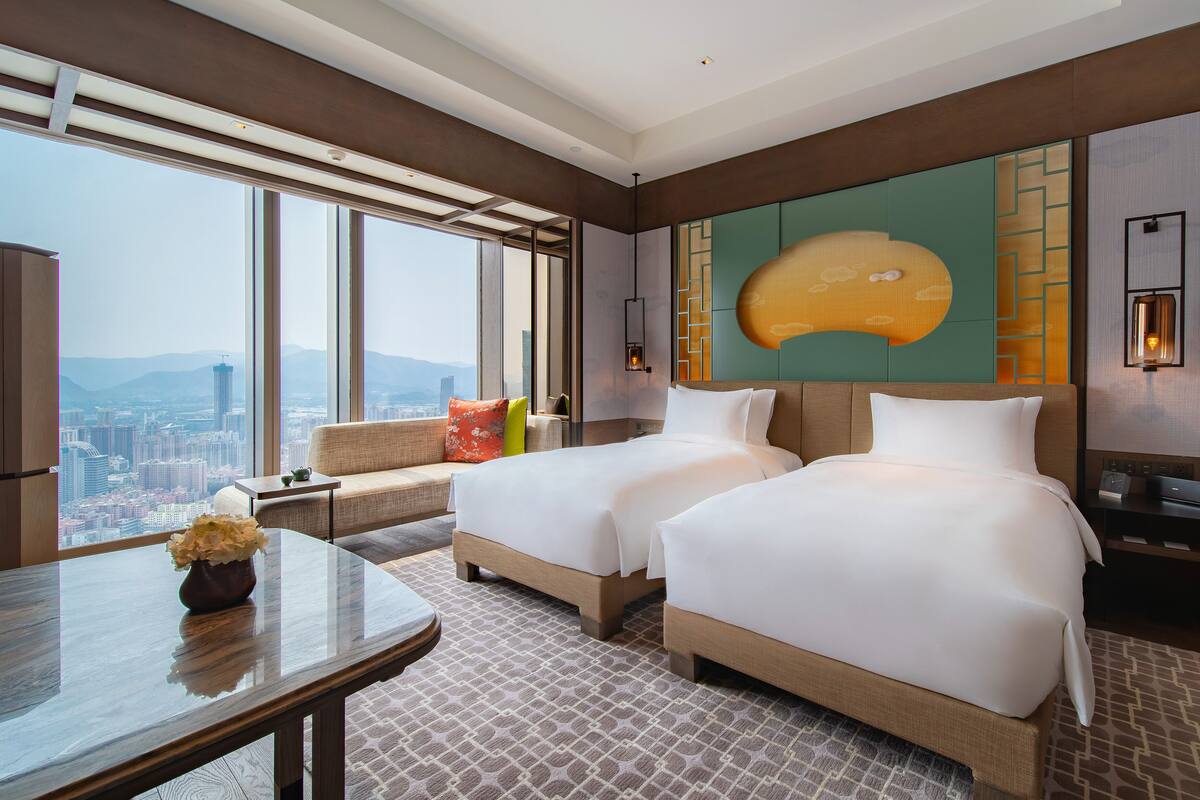 Ein Hotelzimmer mit zwei Betten, einem Schlafsofa, einem Tisch und einem großen Fenster mit Blick auf die Stadt.