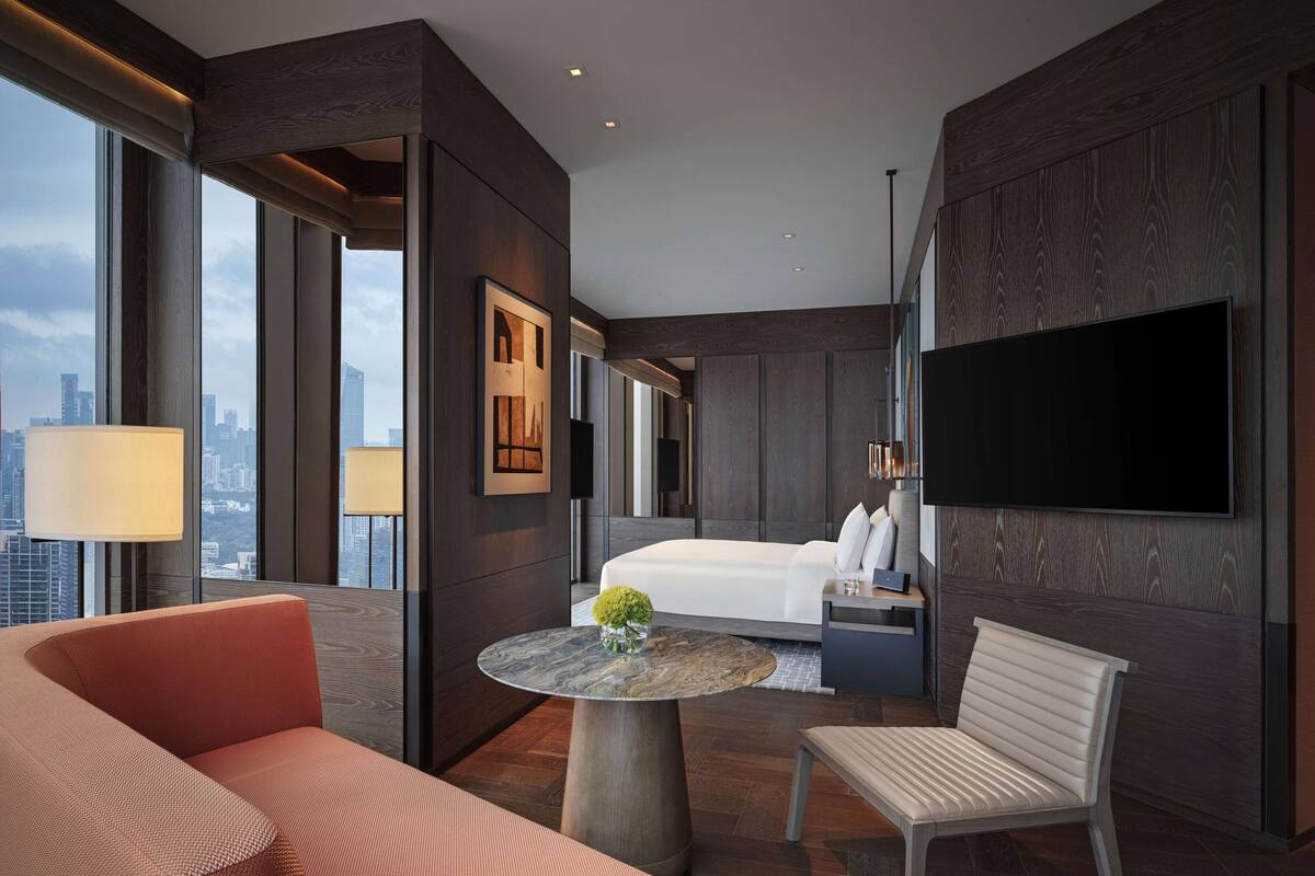 Ein modernes Hotelzimmer mit einem großen Bett, einem an der Wand befestigten Fernseher, einer Sitzecke mit Tisch und Blick auf die Stadt durch große Fenster.