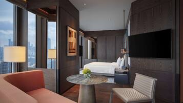 A modern hotel room with a large bed, a TV mounted on the wall, a seating area with a table, and a view of the city through large windows.